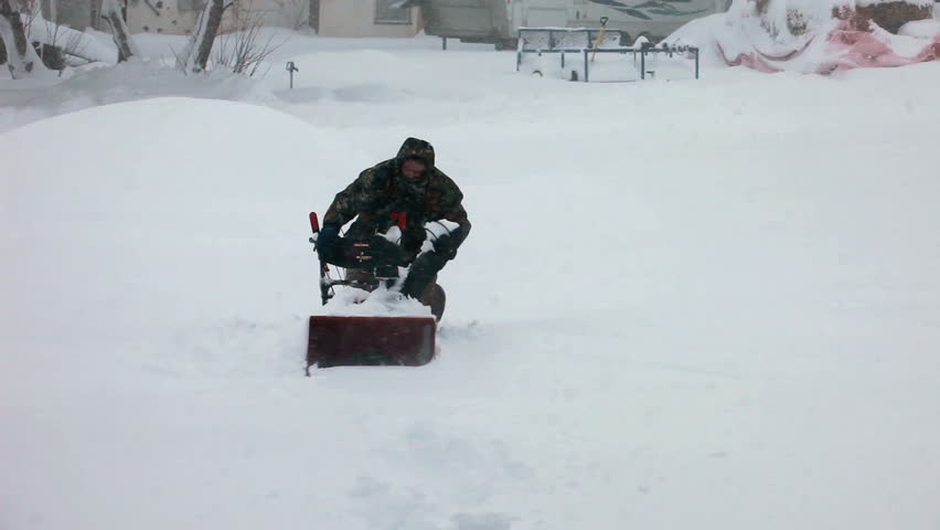 Man starts a snow blower stuck in deep snow blizzard in front of a home. Snow blowing in strong wind in sub freezing weather. Winter time hazard and climate nature.