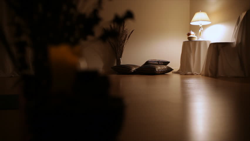 A man dressed in white walks into a room lit only by a small lamp and reposes on the smooth shinny wooden floor to read a book while enjoying his glass of wine.