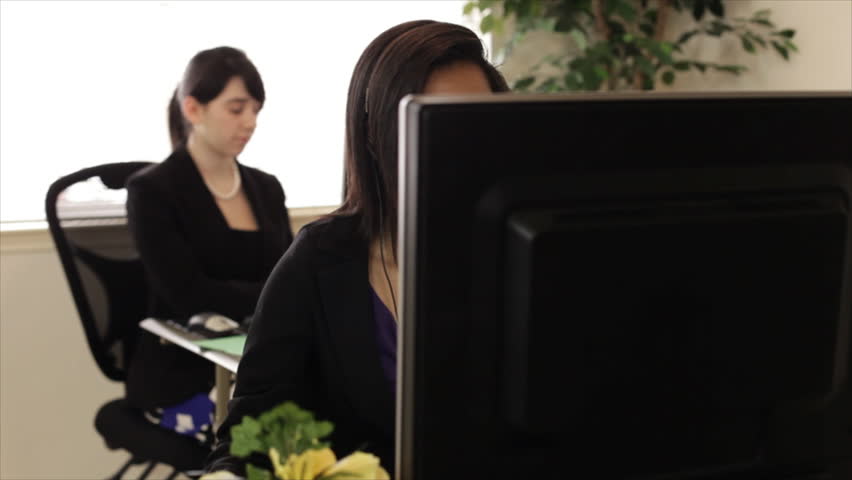 The camera dollies over to reveal a lovely African American girl talking into a headset while working at her computer with soft focus on a pretty coworker typing in the background.