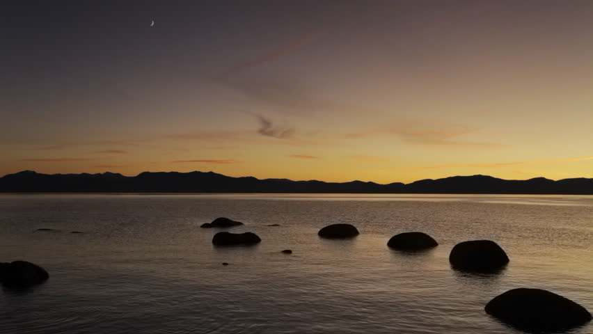 A crescent moon over Lake Tahoe at dusk, with silhouetted, boulders in the foreground and the Sierra Nevada mountains in the background.