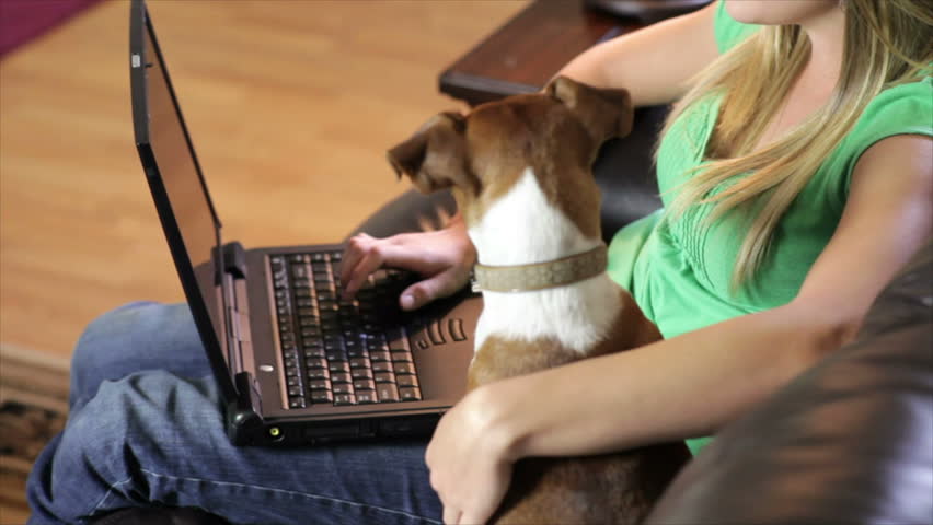 A pretty young woman working on her laptop with a cute little Jack-Rat Terrier dog sitting obediently next to her turns and looks up and smiles for the camera.