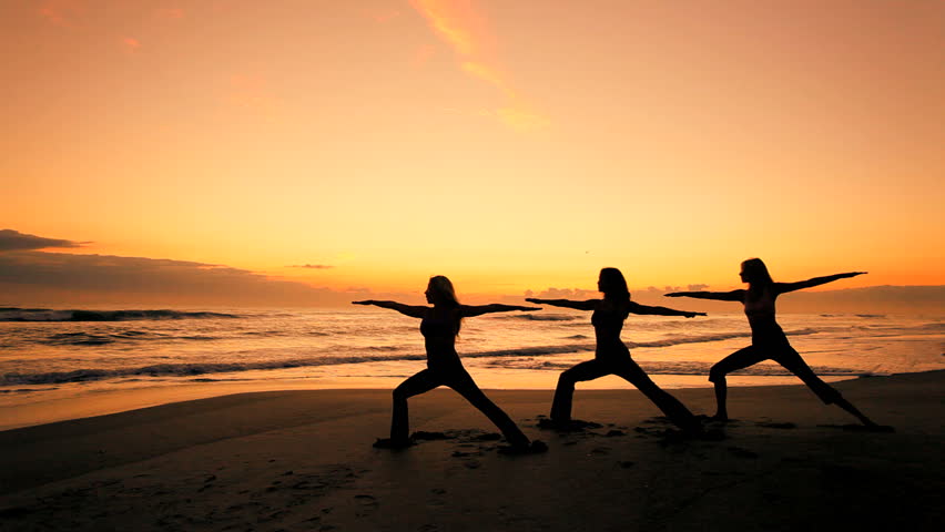 Three slim athletic girls in silhouette practicing a healthy lifestyle with yoga on the beach at sunrise