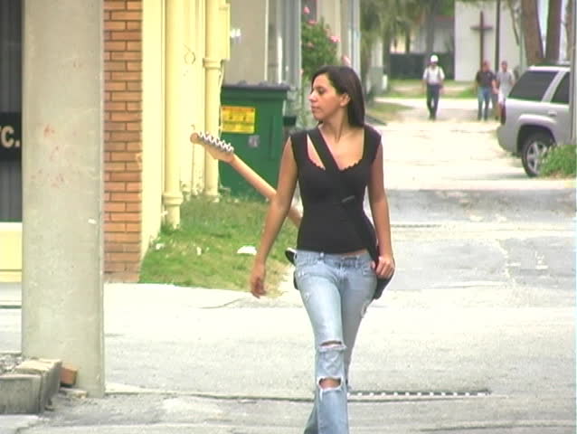 A beautiful young brunette in tattered jeans walks toward the camera with her guitar