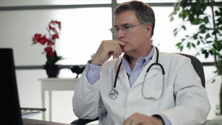 An attractive mature physician working in his office takes off his glasses and gives a welcoming smile to someone off camera. 