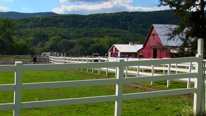 A horse in the background of a white fence and red barn at day in Vermont.