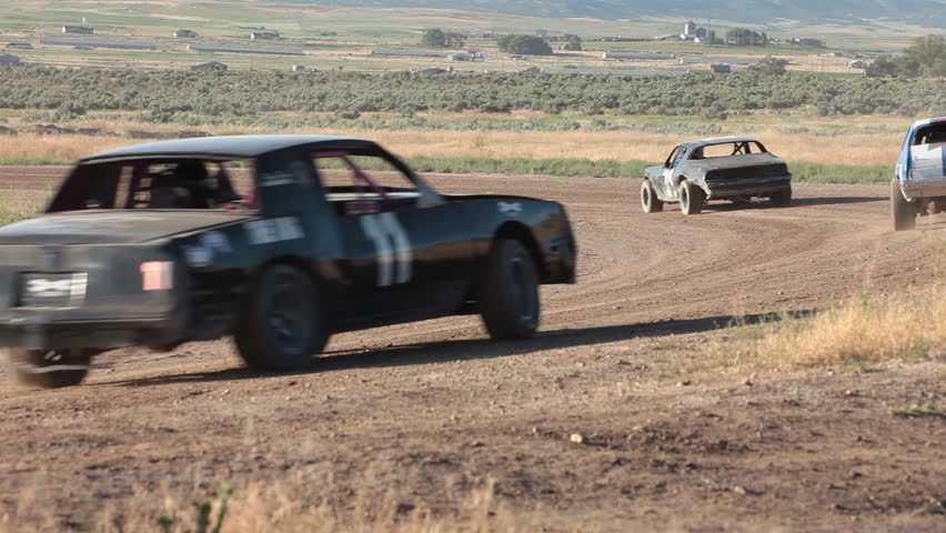 Race stock cars on dirt oval course. Highly modified stock cars driving and racing on a very dirty and dusty track corner. Warmup lap testing the road and track conditions.
