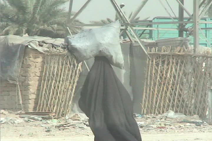 A veiled Muslim woman walks down a street in rural Iraq carrying a bag of goods on her head.