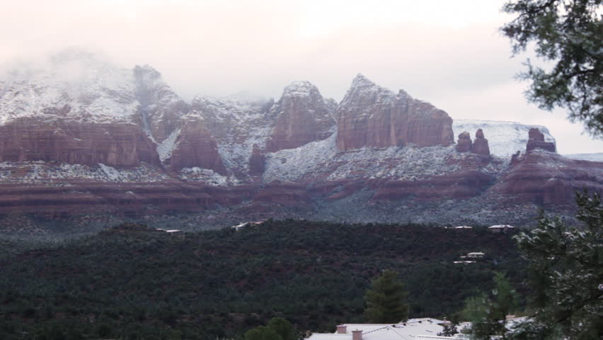 Sunlight shines down through heavy, early-morning clouds covering the mountains of Sedona, Arizona with a light dusting of snow. Wide shot. Juniper trees in foreground.