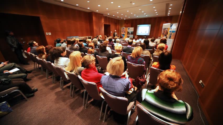 In packed house people listen to performance and observe presentation display on screen