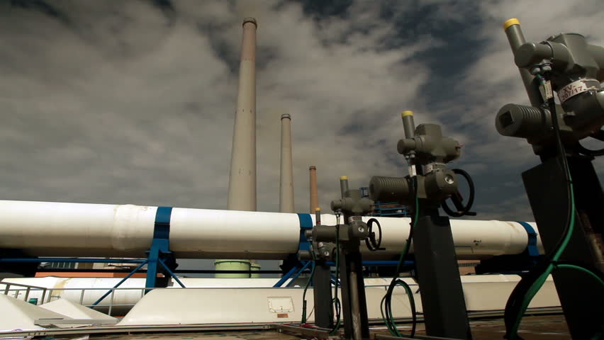Wide dolly along white pipes and valves on the roof of a building with three massive smokestacks in the background at the IDE Ashkelon desalination plant in Israel.