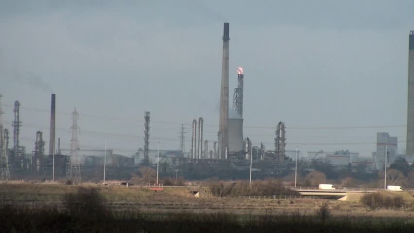 Industrial chimneys belching out smoke into the sky with traffic on the motorway in the foreground.
