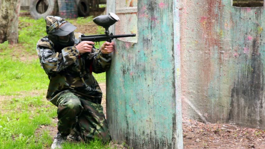 Boy paintball player sits in ambush behind metal fence and looks around
