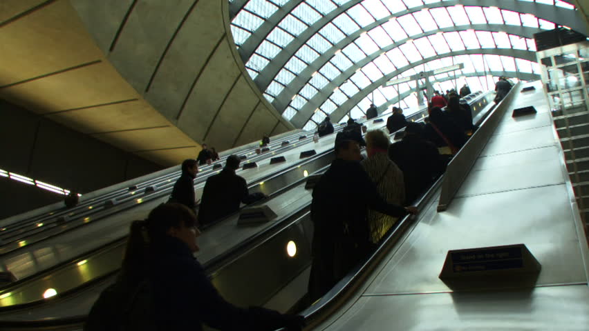 Low angle wide shot of commuters on escalators under glass canopy at Canary Wharf station, the London Underground