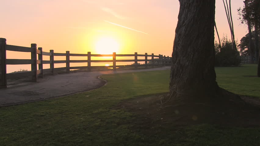 An unrecognizable couple slowly walks along path from left to right as the sun sets on the ocean.