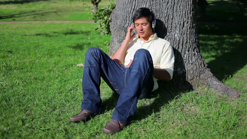Smiling man listening to music while sitting against a tree in a parkland