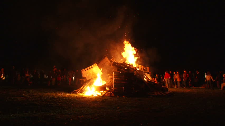 Unrecognizable people watch as huge bonfire gets lit and burns at night, time lapse.
