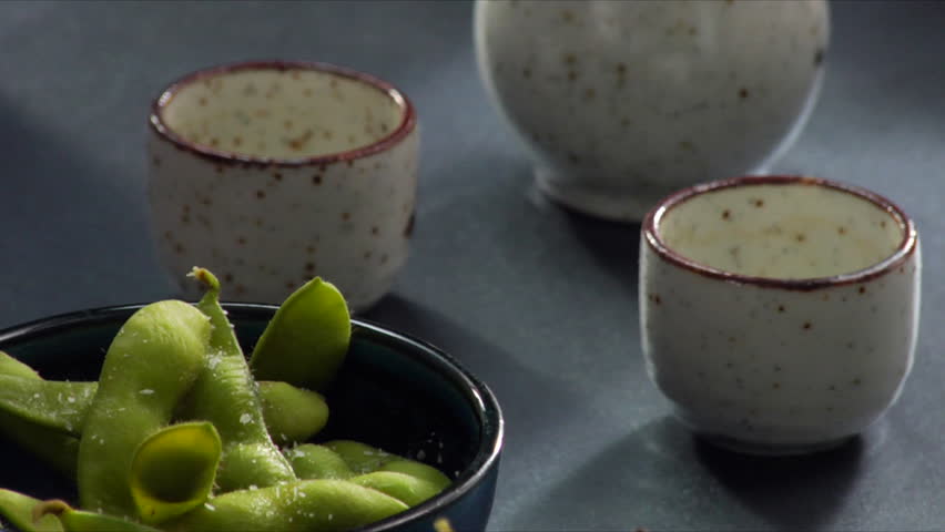 Lock down shot of man pouring hot sake with Edamame (soy beans) in forefront.