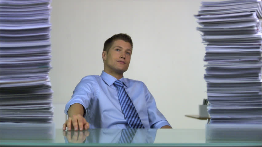 A business man sitting behind a desk, piles of paper decreasing