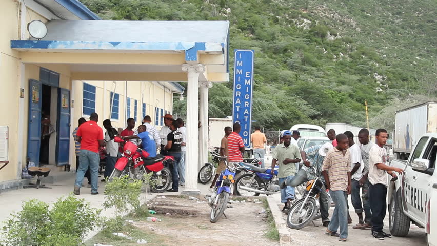 HAITI - JULY 2010:  A group of people stand outside an immigration office, and three men drive by sharing a motorcycle