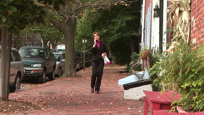 Fall colors surround a woman walking down a brick sidewalk while talking on a cell phone.