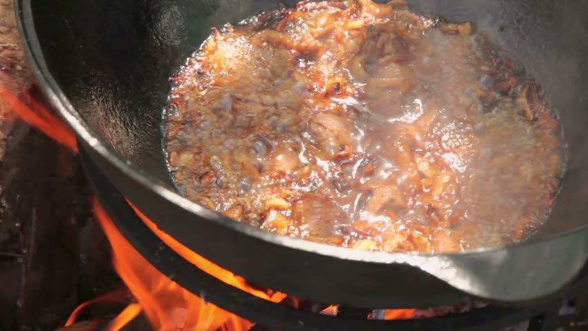 Man mixes broiled onion with meat by colander in pot on campfire