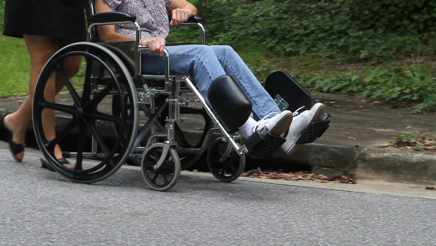 Lower body view of woman caregiver as she pushes disabled senior citizen in a wheelchair down the street.