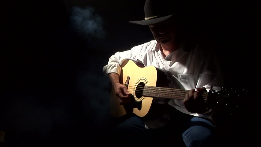 A man in a cowboy hat, playing a guitar spotlighted through the darkness by a smoky beam of light 