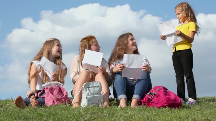 Little girl reads from paper for three girls which sit near on grass at sunny summer day
