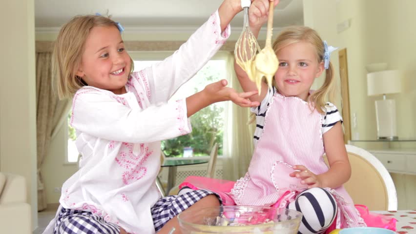 Two girls sitting at kitchen table tasting cake mixture from bowl before putting some on each others noses.
