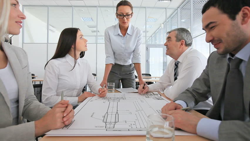 Group of engineers sitting down to table to hold a blueprint discussion under the guidance of their female leader