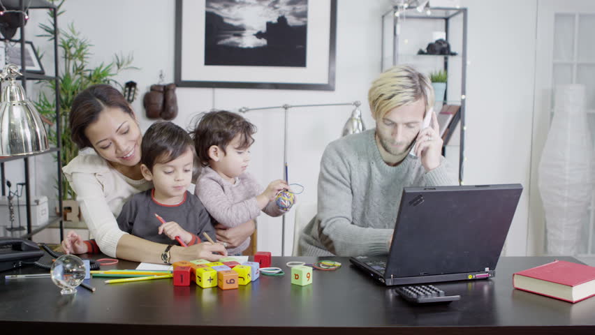 A busy father is working on his laptop and making a telephone call while his wife entertains their children