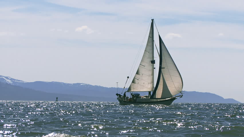 Reverse zoom from a medium framing of a sailboat, either cutter or masthead sloop, under way across the glistening waves of Kachemak Bay in Homer, Alaska