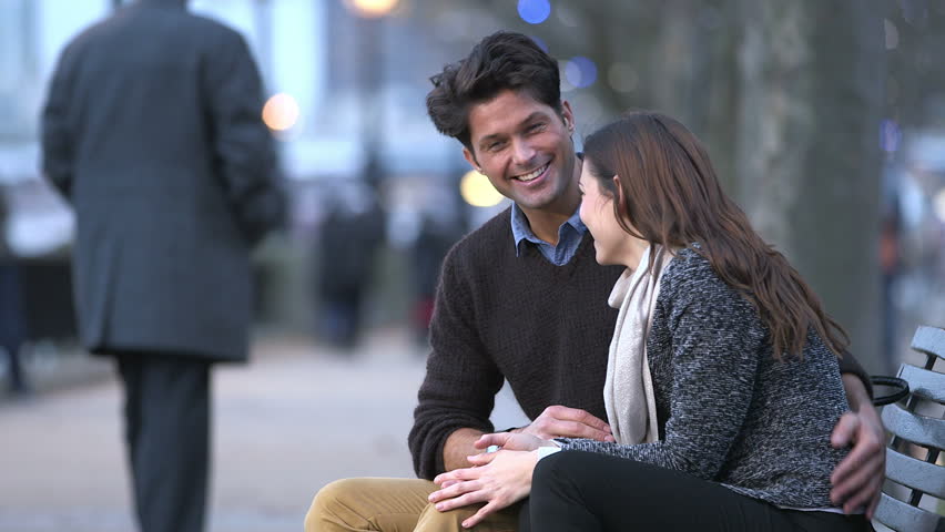 Casually dressed couple enjoy a romantic moment together, sitting on a London bench beside the river thames. The light of the day is just beginning to fade