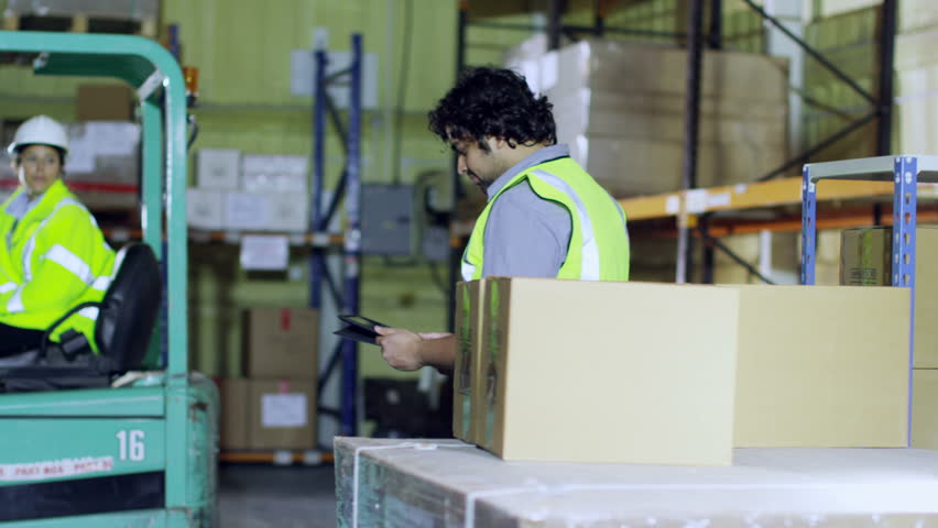 3 workers in high visibility clothing and hard hats are checking stocks and inventory within a warehouse. One man looks into the camera and smiles.