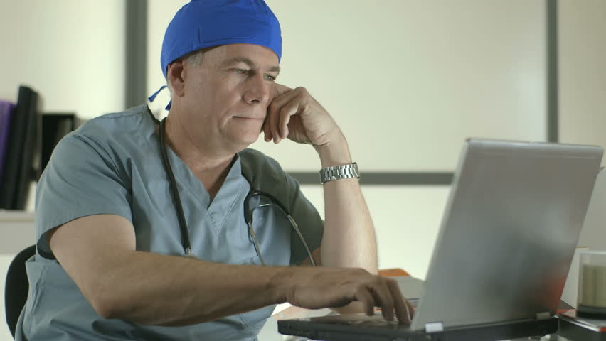 A doctor or surgeon sitting in front of a laptop shows fatigue as he removes his stethoscope and cap as if calling it a day. 