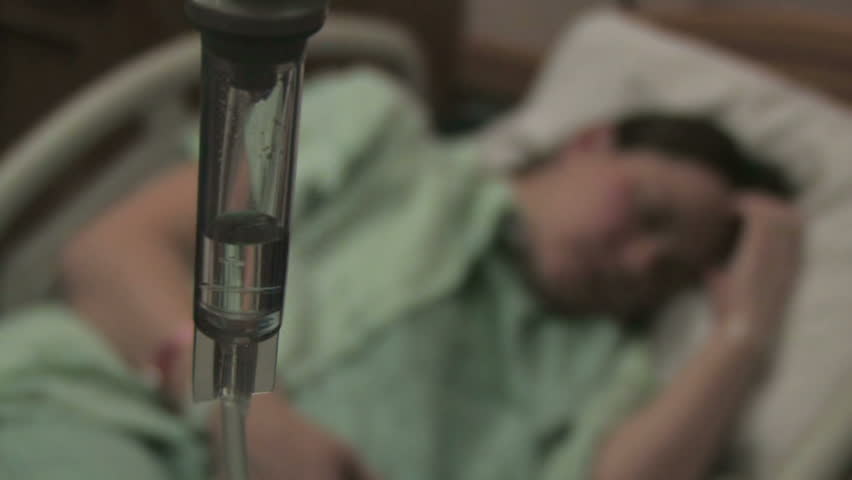 A sick woman lays in a hospital bed behind an IV drip chamber.