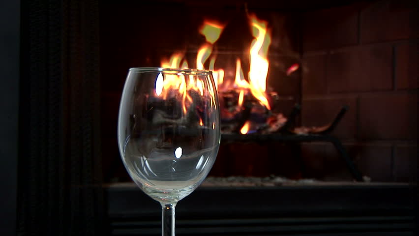 Radial dolly shot of a woman pours a glass of red wine with a fire burning in a fireplace in the background.