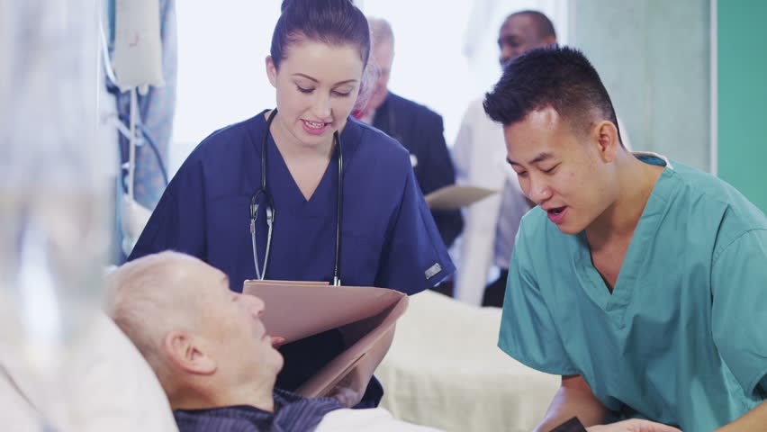 One male and one female nurse attend to an elderly male patient, checking his blood pressure and chatting with him. In the background a nurse and consultant specialist are discussing another patient.
