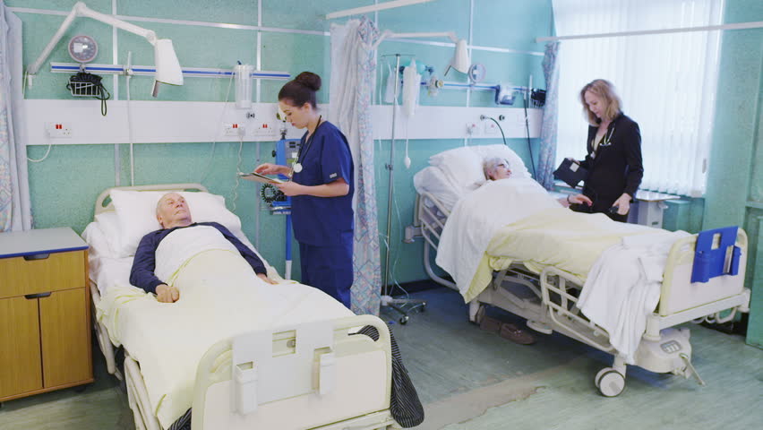 Doctors and nurses on a UK hospital ward are busy taking care of two elderly patients. In slow motion.