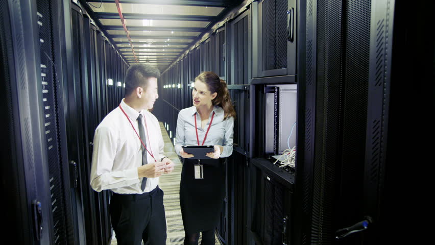 Three people of mixed ethnicity are working in a data center with rows of server racks and super computers. They are walking up and down and checking all of the equipment. 