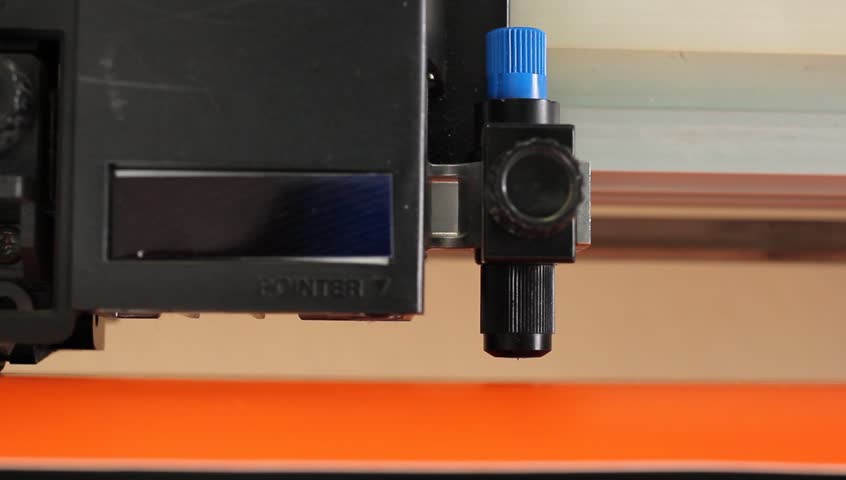 Cutting plotters use knives to cut the letters on orange vinyl film.  Close-up view from below
