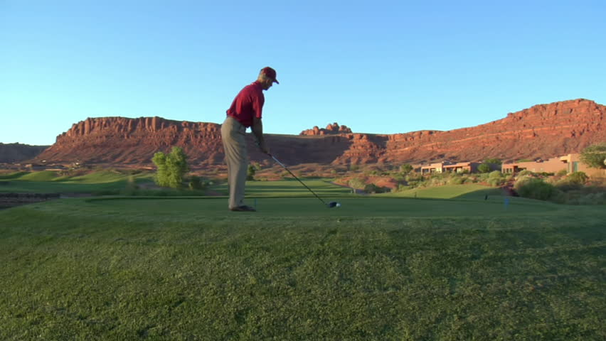 steadicam shot of man teeing off on golf course with sunset and red rock cliffs
