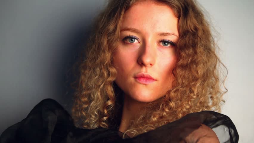 Young model with curly hair stands near wall wrapped in black shawl