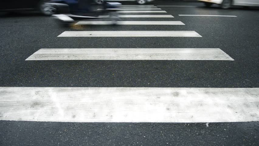 Different types of transport crossing smooth road. One-way traffic. Cars, buses, scooters and bikes in motion. Focus on white lines of pedestrian crossing and tires of cars. Busy street life.