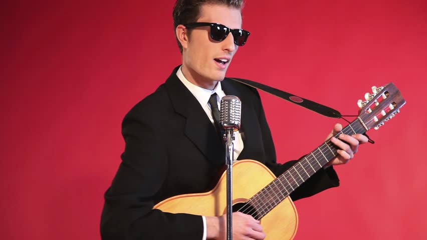 Retro fifties rock and roll singer playing acoustic guitar. Wearing black suit and tie and sunglasses. Studio shot. (No audio).