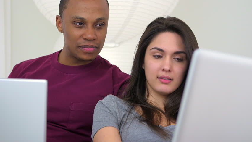 African American and Caucasian couple using laptop computers on couch
