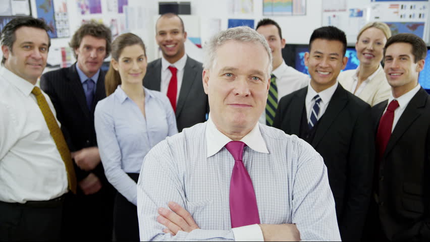 Portrait of a mature businessman who is standing in front of the rest of his diverse and multi ethnic business team. They all look into the camera and smile. 
