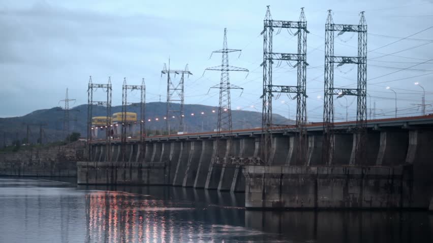Trucks ride by dam of Hydroelectric power station on river at evening, pillars with high-voltage wires