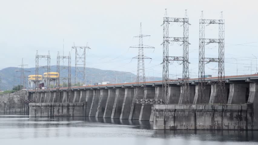 Trucks ride by dam of Hydroelectric power station on river, pillars with high-voltage wires