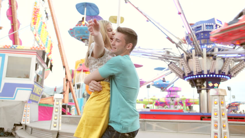 Joyful young couple visiting an amusement park arcade with man picking up girl with balloons and turning around smiling and having fun.